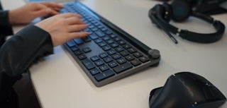 Hands on a JLAB Epic Wireless Keyboard with JLAB Headset in background and JLAB wireless mouse in foreground